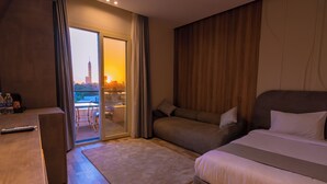 A hotel room with a bed, a sofa, a desk with a remote control, and a view of a cityscape through a large window. - Elevana Boutique hotel (Cairo)