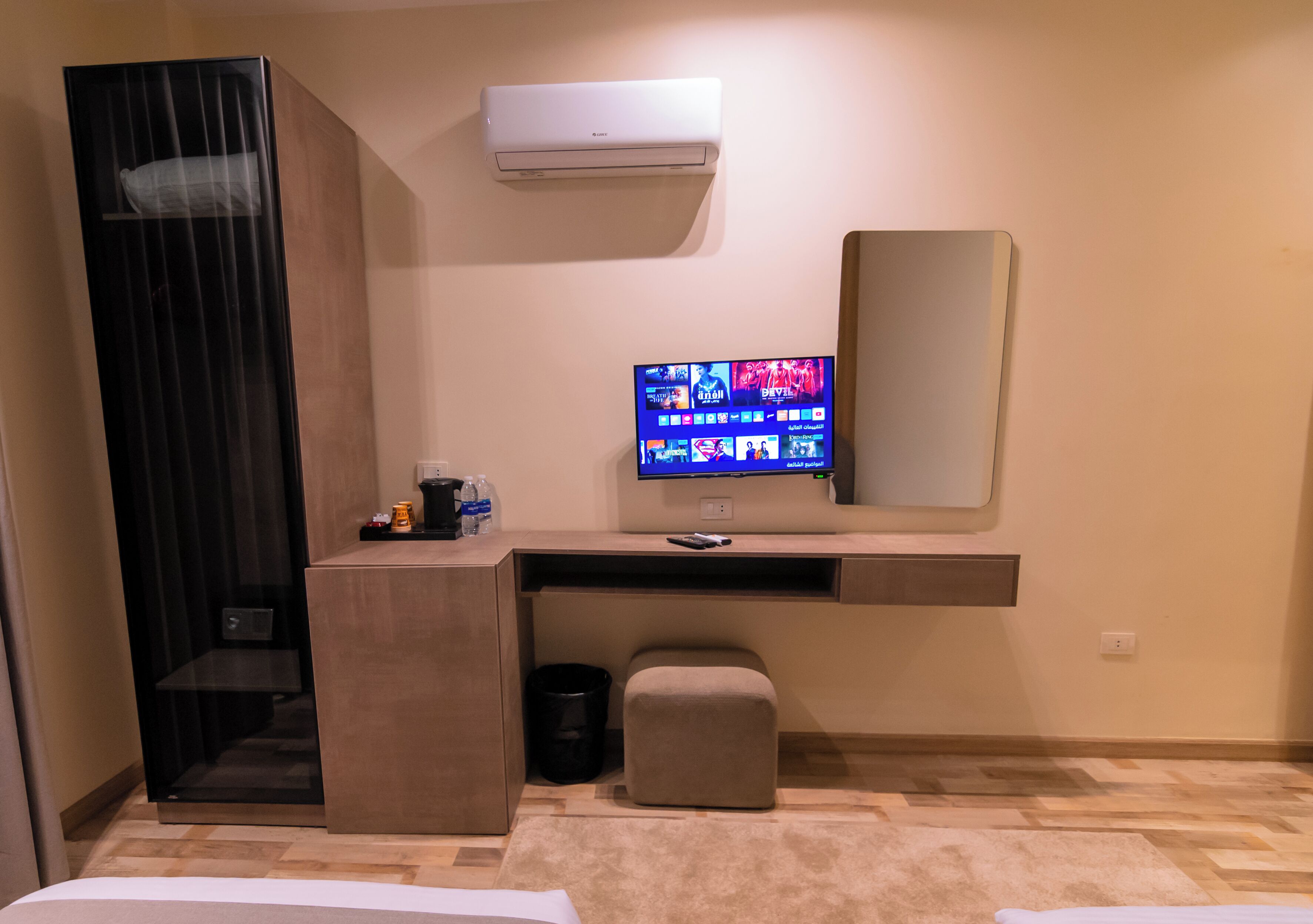 Un dormitorio moderno con una cama, un escritorio con ordenador, un aire acondicionado y un pequeño reposapiés.