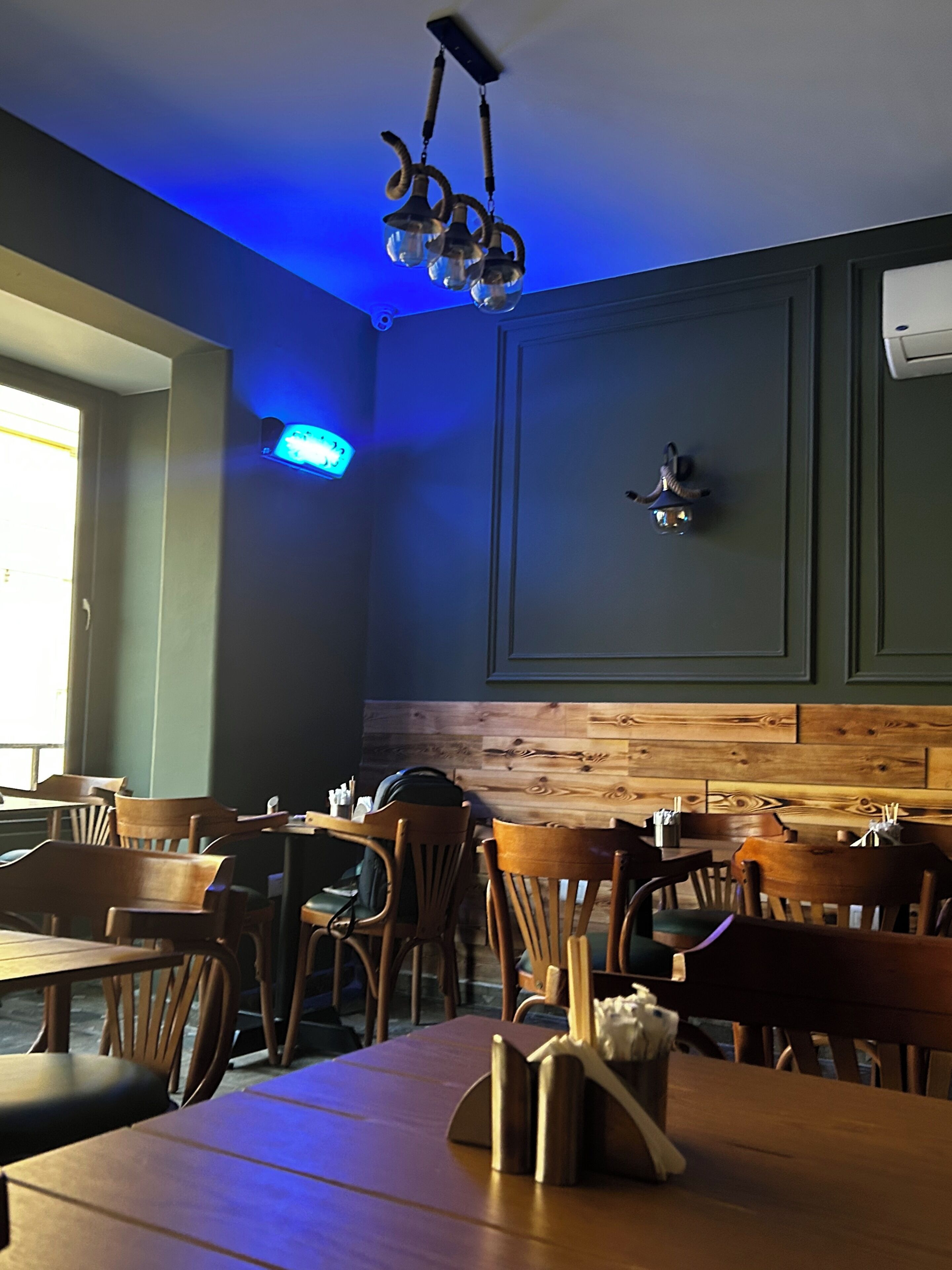 A restaurant interior with wooden tables and chairs, a blue-lit ceiling, and a wooden accent wall.