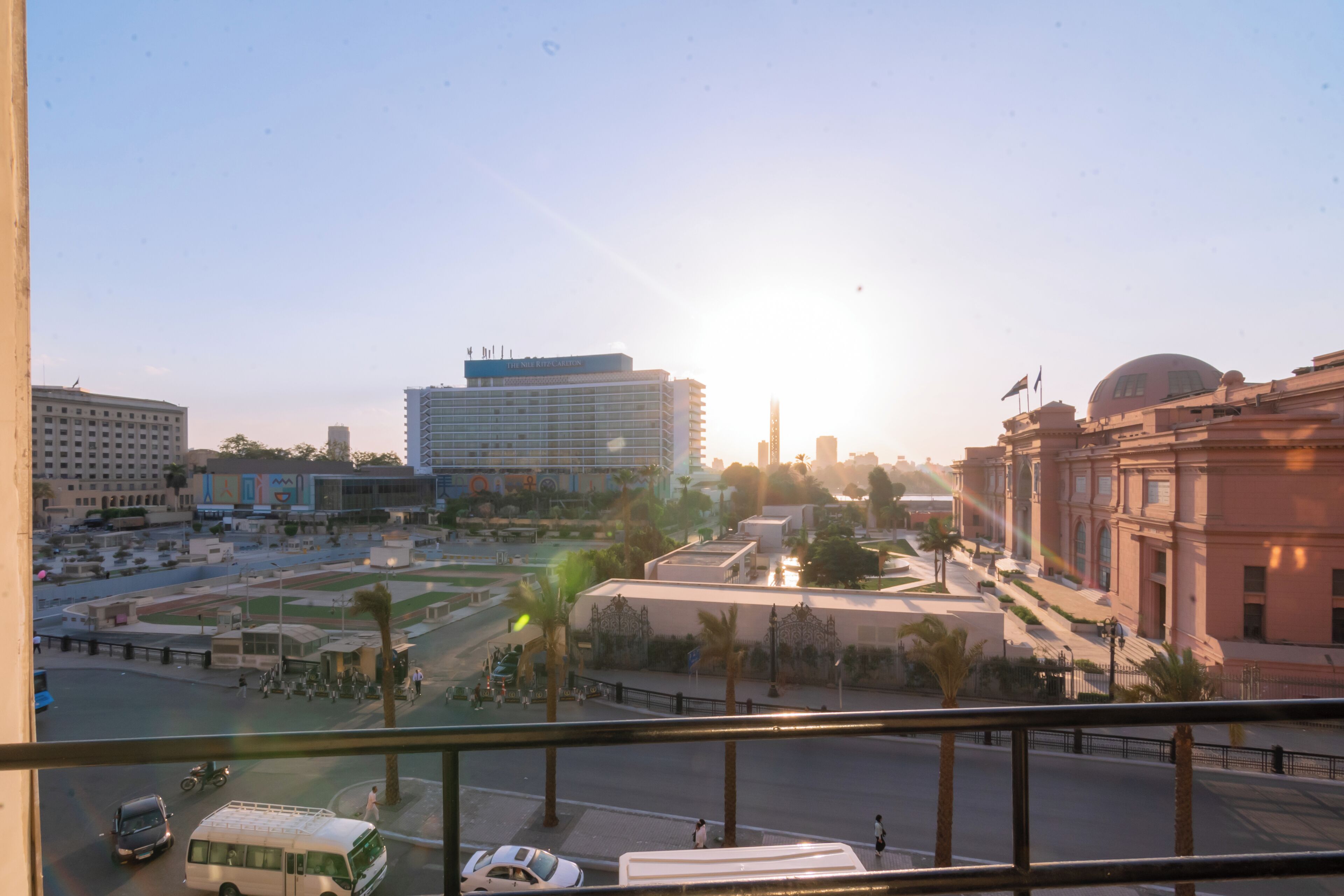 A cityscape with a prominent building featuring a dome and flags, surrounded by palm trees and a clear sky.
