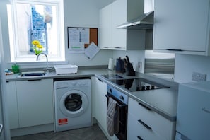 A compact kitchen with a washing machine, a stove, and a window with a view of a stone building.