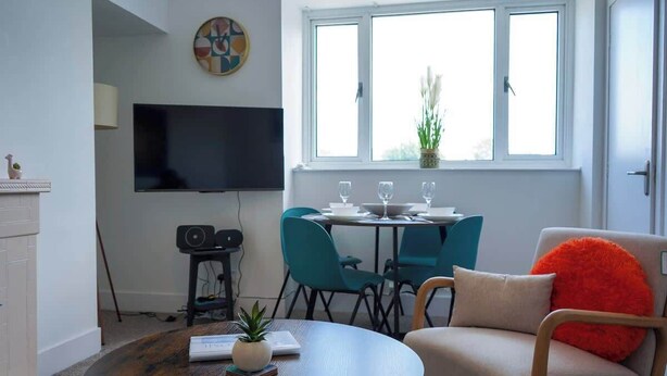 Een woonkamer met een ronde salontafel, een bank met een rood kussen, een aan de muur bevestigde televisie, en een eethoek met stoelen en een tafel.