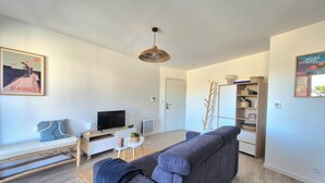 A modern living room with a grey sofa, a TV, a bookshelf, and a hanging light fixture. - Beautiful apartment with balcony and garden (Saint-Coulomb)