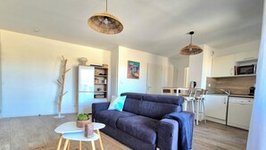 A modern living room with a sofa, a small table, and a rug. There is a kitchen area with cabinets and a microwave. - Beautiful apartment with balcony and garden (Saint-Coulomb)