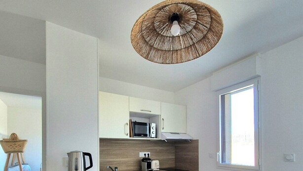 A modern kitchen with white cabinets, a wooden dining table, and a woven ceiling light fixture. - Beautiful apartment with balcony and garden (Saint-Coulomb)
