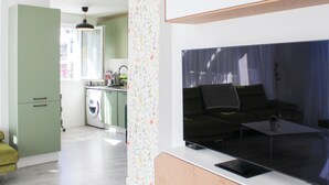 Ein modernes Wohnzimmer mit großem Fenster, einer Couch, einem Couchtisch und einer Küchenzeile inklusive Waschmaschine.