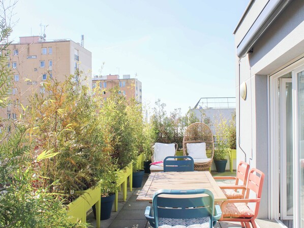 Une terrasse extérieure avec une table et des chaises, entourée de plantes en pot et offrant une vue sur des immeubles d’habitation.