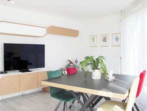 Un coin repas moderne avec un téléviseur à écran plat, un meuble en bois, une table à manger et des chaises. Des reproductions botaniques encadrées ornent le mur, et une plante en pot est posée sur la table.