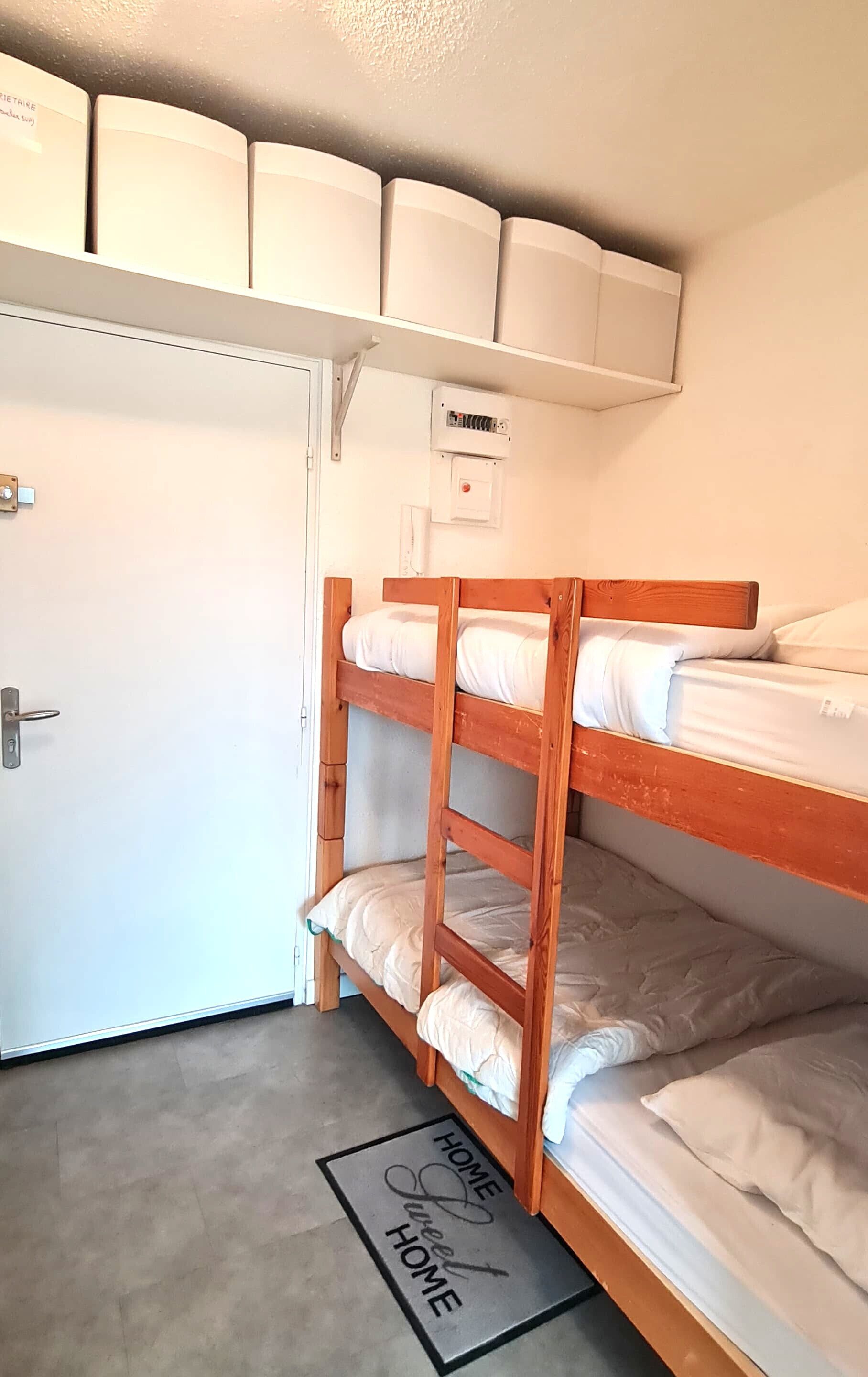 Une chambre avec un lit superposé, une porte blanche, une étagère au-dessus du lit avec des boîtes de rangement, un petit tableau électrique et un paillasson avec l'inscription « HOME Sweet HOME ».