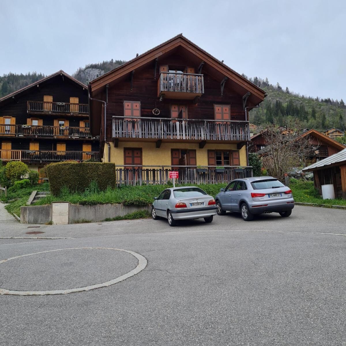 A multi-story wooden building with balconies, parked cars, and a mountainous backdrop.