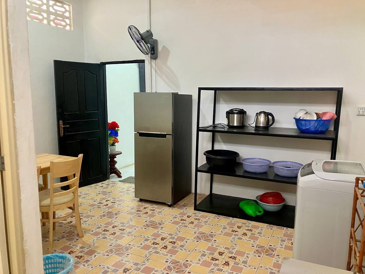 A compact kitchen with a refrigerator, a shelf with kitchenware, a fan, and a tiled floor.