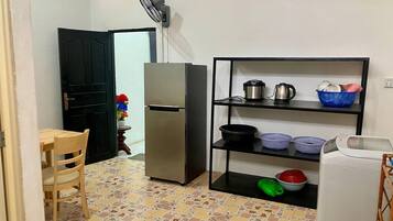 A compact kitchen with a refrigerator, a shelf with kitchenware, a fan, and a tiled floor.