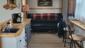 A compact kitchen with a sink, coffee maker, and bar stools. A black leather sofa is positioned in the center with a plaid blanket.