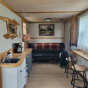 A compact kitchen with a sink, coffee maker, and bar stools. A black leather sofa is positioned in the center with a plaid blanket.