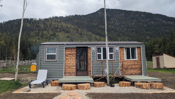 A small cabin with a wooden exterior, a front door, windows, and a porch area with a bench and wooden stools.