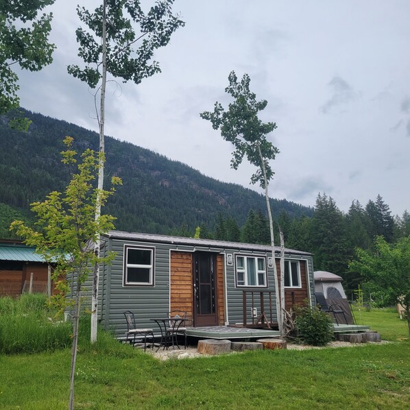 A small cabin with a front porch, a wooden door, and a window. - Tiny House with stunning mountain views, steps from Arrow lake. Pet- friendly! (Nakusp)
