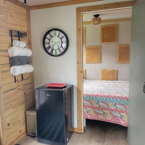 Una habitación con una cama, un reloj en la pared, un refrigerador pequeño negro y una pared de madera con toallas.