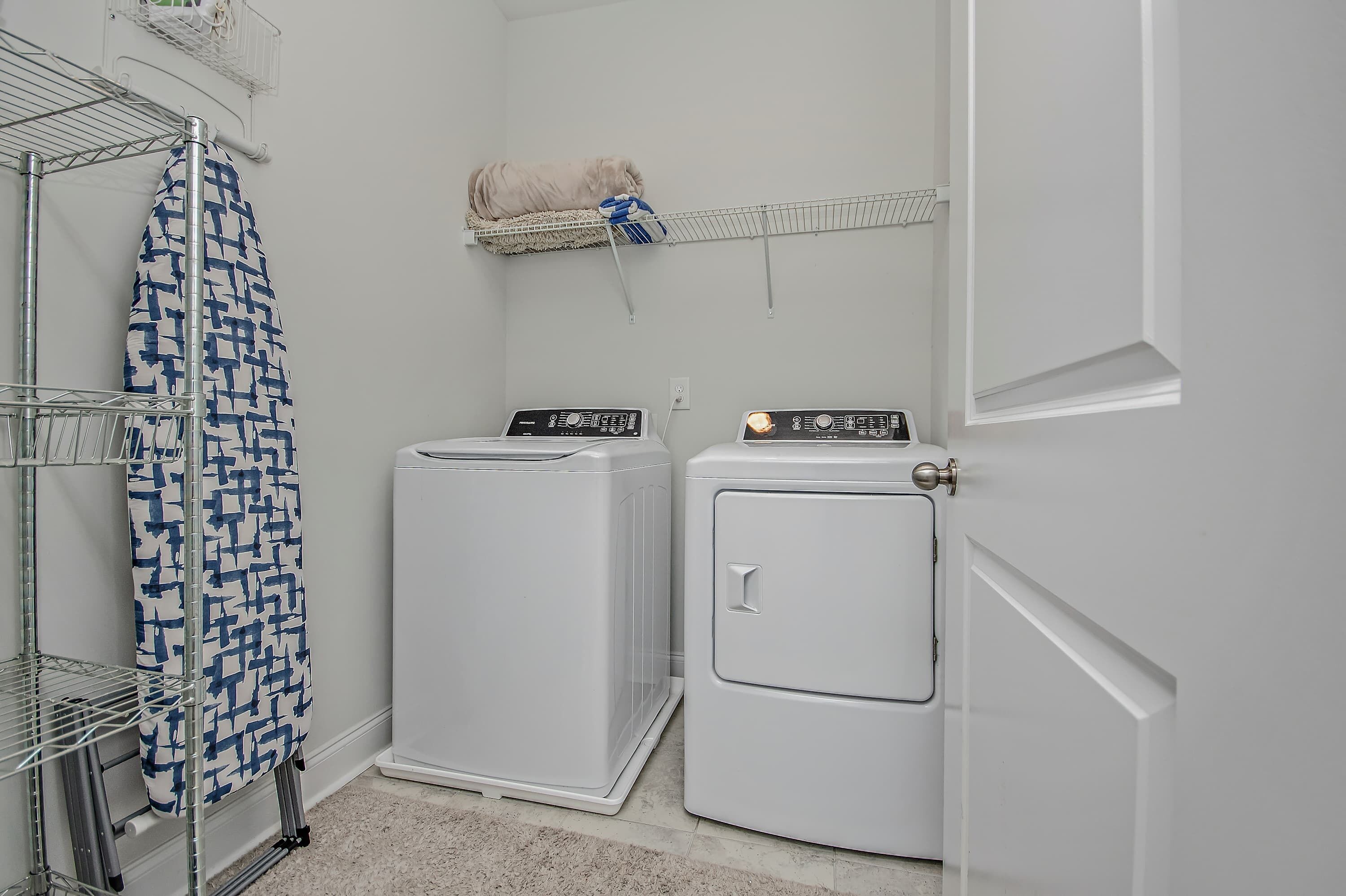 A laundry area with two white washing machines, a drying rack with a patterned cloth, and a metal shelf with a rolled-up towel.