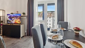 Een eethoek met een tafel gedekt voor twee personen, een televisie op een kast, en een uitzicht op gebouwen door een raam.