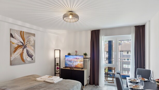 Een moderne hotelkamer met een bed, een televisie, een eettafel met stoelen en een balkon met uitzicht op gebouwen.