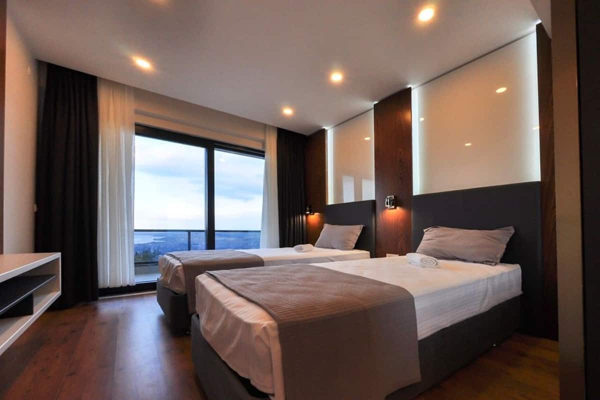 Ein modernes Hotelzimmer mit zwei Betten, einem großen Fenster und Stadtblick sowie einem kleinen Schreibtisch.