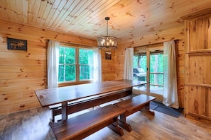 Interior de una cabaña de madera con una gran mesa de comedor, dos bancos, una araña y ventanas amplias con cortinas.