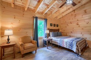 Une chambre confortable dans un chalet, avec un plafond en bois, un lit recouvert d’une couverture matelassée, un fauteuil en cuir, une petite table avec une lampe et une fenêtre avec des rideaux.