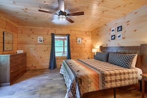 Un dormitorio con paneles de madera, una cama, cómoda, ventilador de techo y decoración en la pared.