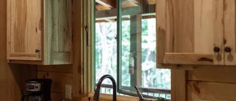 Una cocina con armarios de madera, un lavamanos, una cafetera y un ventanal con vista a árboles.