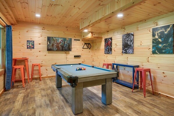 A pool table in a room with wooden walls and ceiling, surrounded by stools and paintings.