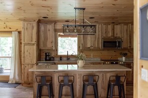 A wooden cabin kitchen with a central island, bar stools, a hanging light fixture, and a window with a view. - Night Song Lodge | Hocking Hills, New, Sleeps 16 (Logan)