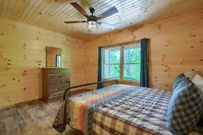 A wooden cabin bedroom with a bed, dresser, and window with curtains.