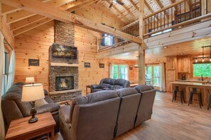 A cozy cabin interior with a stone fireplace, wooden walls, a comfortable seating area, and a kitchen with a bar.