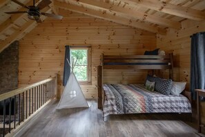 A wooden cabin room with a bunk bed, a window with a view of greenery, a wooden ceiling, and a small window with a curtain. - Night Song Lodge | Hocking Hills, New, Sleeps 16 (Logan)