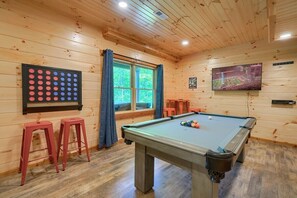 A pool table with stools, a dartboard, and a TV in a wooden room with a window.