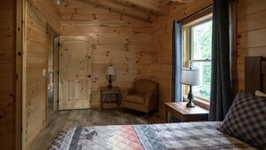 Ein Zimmer in einer Holzhütte mit einem Bett, einem Sessel, einem kleinen Tisch und einem Fenster mit Vorhängen.