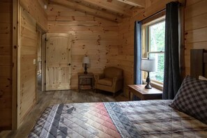 Een houten kamer met een bed, een stoel, een klein tafeltje en een raam met gordijnen.
