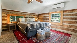 A bedroom with a wooden log wall, a bed with a striped blanket, a red and white rug, and a ceiling fan.