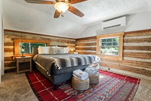 A bedroom with a wooden log wall, a bed with a striped blanket, a red and white rug, and a ceiling fan.