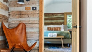 A rustic room with a wooden wall, a single bed with a green quilt, a wooden chair, and a small table.