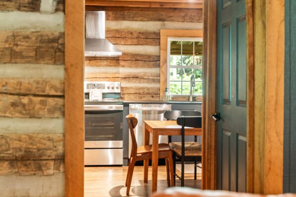 A rustic log cabin interior with a wooden dining table, chairs, a stove, and a window with a view of greenery. - Lodge 1880 | Hocking Hills, Historic Log Cabin (Rockbridge)