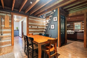 A rustic log cabin interior with a wooden dining table, chairs, and a cozy seating area with a bench and cushions.