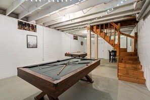Ein Billardtisch mit Billardstöcken, eine Dartscheibe und eine Shuffleboard-Spielfläche in einem Raum mit weißen Backsteinwänden, einer Holztreppe und Lichterketten an der Decke.