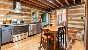 A log cabin interior with a kitchen featuring stainless steel appliances, a wooden dining table, and a large window.