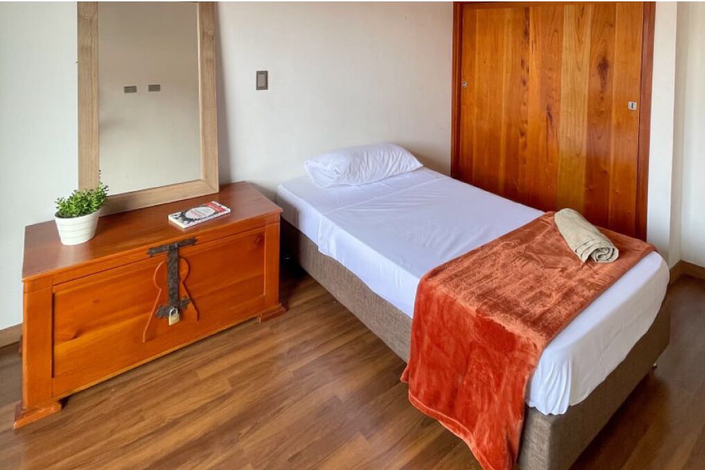 Une chambre avec une commode en bois, un lit recouvert d’un drap blanc et d’une couverture orange, un miroir et une armoire en bois.