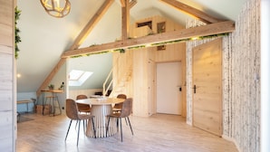 A spacious loft with a high ceiling, exposed wooden beams, a round dining table with chairs, and a staircase leading to an upper level. - La Cabane - deux chambres et grande terrasse  ⭐️⭐️⭐️ (Metz)
