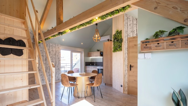 A modern kitchen with wooden beams, a dining area with a round table and chairs, and a staircase with a wooden railing. - La Cabane - deux chambres et grande terrasse  ⭐️⭐️⭐️ (Metz)