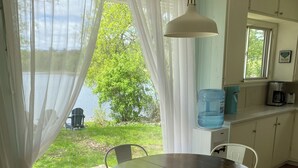 A dining area with a round table and four chairs, a large window with sheer curtains, and a view of a lake and greenery outside.