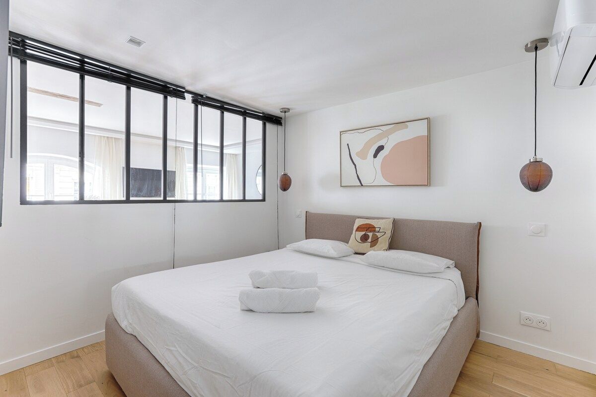 A modern bedroom with a large bed, a window with blinds, a wall-mounted air conditioner, and a single pendant light.