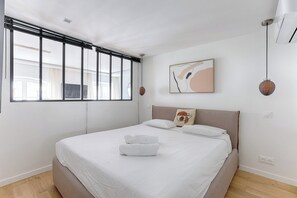 Un dormitorio moderno con una cama grande, una ventana con persianas, un aire acondicionado de pared y una lámpara colgante.